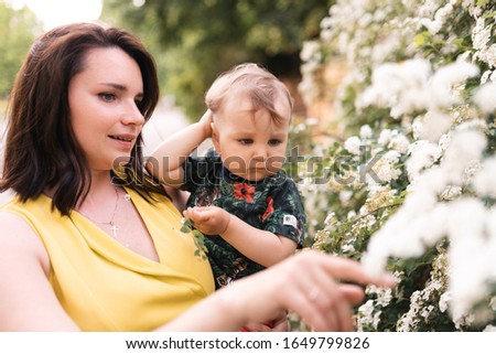 Mother playing and talking to baby child boy with flower blossoming tree in the background - Young mom in green dress and her baby in a green park having fun - Happy smiling people