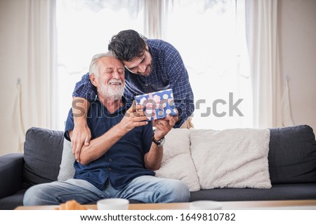 Happy people family concept - Old senior old man opens the gift box the son gave. Young man hugs his elderly father. Two men sitting on the sofa in the house.
