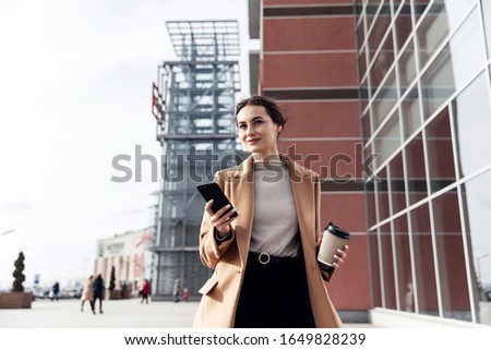 Young cousian woman with smartphone standing against street blurred building background. Fashion business photo of beautiful girl in black casual suite with phone and cup of coffee