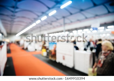 Defocused view of large hall with silhouettes of people visiting the exhibition fair with multiple booths