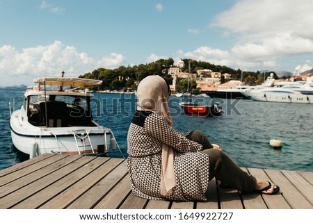 woman with hijab sitting on deck by the sea.  Female tourist exploring Cavtat. She is sitting turned back and looking in the old city. 