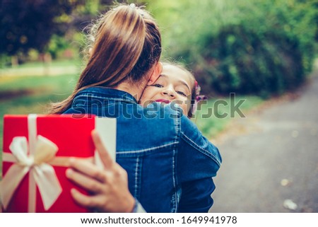 Two school friends hugging, emotional meeting in the park. Happy little girl gives her smiling friend a gift in a red box, making a surprise. People, children, holidays, friends and friendship concept