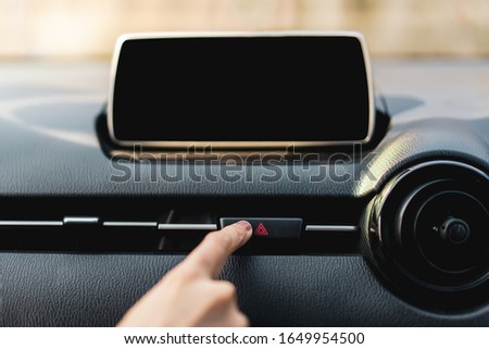 Driver pressing on hazard button on a car dashboard close up with empty infotainment screen (entertainment screen inside a car). Woman pushing on red hazard button on car dashboard with empty monitor.