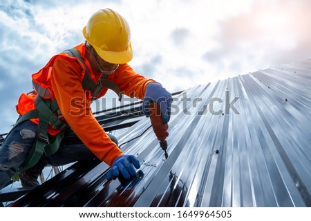 Close up photo of Professional and qualified roofer in protective uniform wear use electric drill to install the metal sheet on the new roof of new modern building construction.