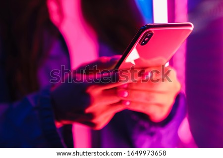 Cropped photo of young woman posing over neon lights using mobile phone.
