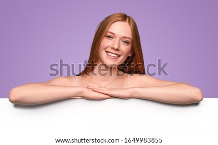 Optimistic ginger female smiling and looking at camera while standing behind blank poster and advertising beauty industry against violet background