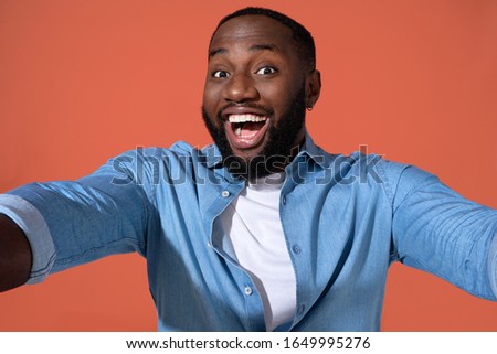 Cheerful man taking selfie on the phone. Photo of african man in casual outfit on coral background.