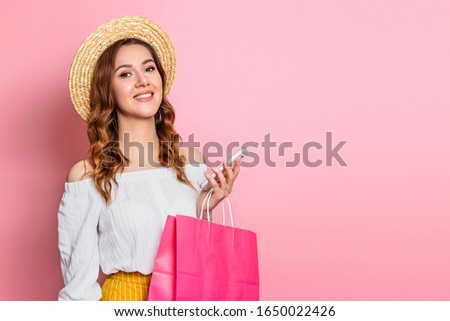 Young woman in a straw hat and a white summer dress with a mobile phone and shopping bags smiling on a pink background, copy space for text, web banner. Girl makes online shopping, photo in the studio