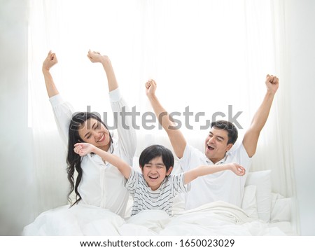 Happy family wake up in morning stretching hand rise up to the air while sitting on bed in bedroom with big window in background.Caucasian young man with asian woman and little boy exercise on bed. Royalty-Free Stock Photo #1650023029