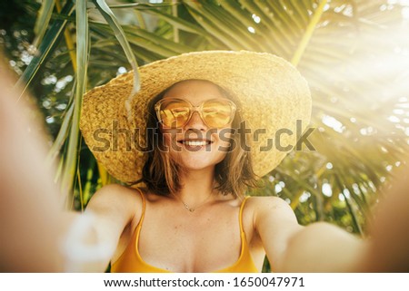 Sunny picture of young woman resing on beach under palm trees on island. Take slefie by holding camera with both hands and smile. Stylish modern female model wear hat and sunglasses