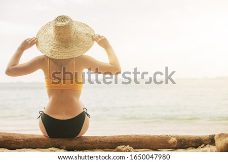 PIcture of young woman with attractive body sit on ocean coast and look at sea or water. Wear swimsuit and hold edge of her straw hat. Vacation abroad and rest mood on tropical island