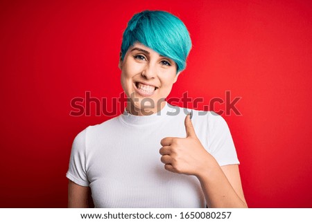 Young beautiful woman with blue fashion hair wearing casual t-shirt over red background doing happy thumbs up gesture with hand. Approving expression looking at the camera showing success.