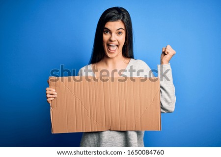 Young beautiful brunette activist woman holding blank cardboard banner protesting screaming proud and celebrating victory and success very excited, cheering emotion