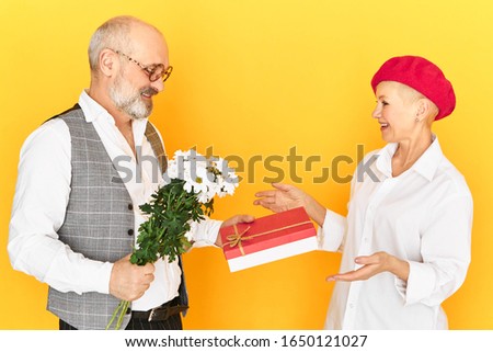 Isolated image of beautiful European mature lady receicing box of candy and field flowers from her elderly boyfriend in elegant clothes and eyewear. Shy senior male making birthday gift to his wife