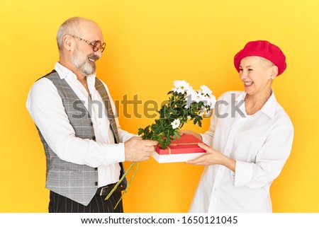 Horizontal image of shy awkward grandfather wuth gray beard holding flowers and box of present congratulating his mature girlfriend on her birthday. Cute happy elderly couple on first date