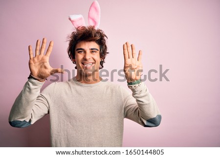 Young handsome man holding easter rabbit ears standing over isolated pink background showing and pointing up with fingers number nine while smiling confident and happy.
