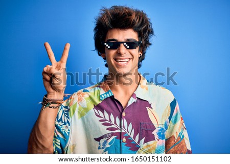Young handsome man wearing thug life glasses standing over isolated blue background smiling with happy face winking at the camera doing victory sign. Number two.