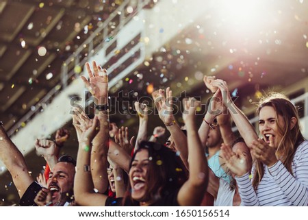Sports fans clapping hands in stands with falling confetti.  Football team supporters cheering in stadium.