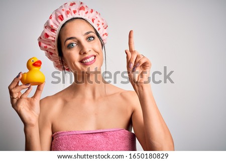Young brunette woman with blue eyes wearing bath towel and shower cap holding duck toy surprised with an idea or question pointing finger with happy face, number one