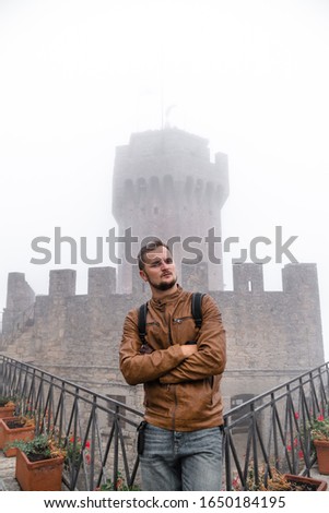 Handsome man wearing leather jacket and backpack. Fortification, old castle on background. Journey. Mystical atmosphere, fog, white haze, mist. Vertical photo. Second Tower. San Marino, Italy
