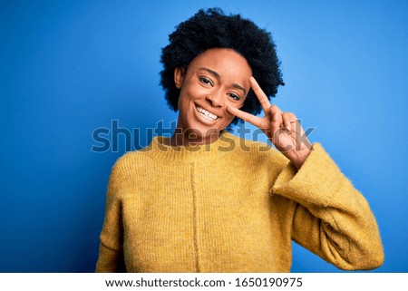 Young beautiful African American afro woman with curly hair wearing yellow casual sweater Doing peace symbol with fingers over face, smiling cheerful showing victory