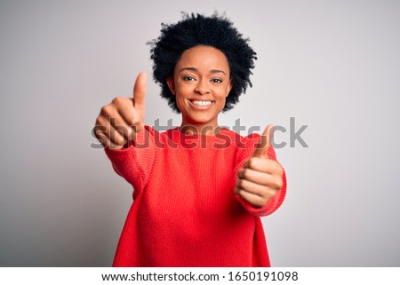 Young beautiful African American afro woman with curly hair wearing red casual sweater approving doing positive gesture with hand, thumbs up smiling and happy for success. Winner gesture.