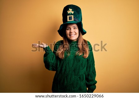 Young beautiful brunette woman wearing green hat on st patricks day celebration smiling cheerful presenting and pointing with palm of hand looking at the camera.