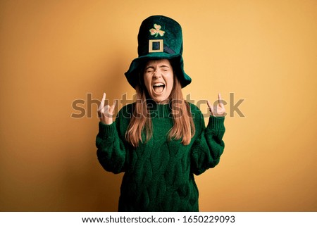 Young beautiful brunette woman wearing green hat on st patricks day celebration shouting with crazy expression doing rock symbol with hands up. Music star. Heavy concept.