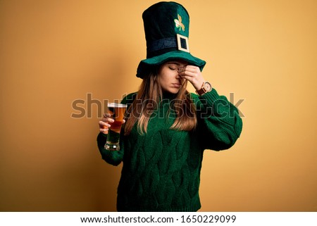 Young beautiful woman wearing green hat drinking glass of beer on saint patricks day tired rubbing nose and eyes feeling fatigue and headache. Stress and frustration concept.