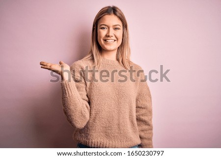 Young beautiful blonde woman wearing winter wool sweater over pink isolated background smiling cheerful presenting and pointing with palm of hand looking at the camera.
