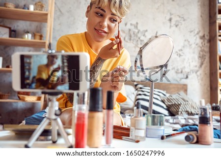 Young woman make-up blogger influencer sitting at stylish urban apartment recording vlog on smartphone camera looking at mirror reflection applying eyeshadow smiling joyful