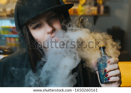 Young pretty woman in cap smoke an electronic cigarette at the vape shop. Hip-hop style. Closeup.