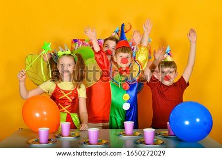 Four children with a clown at the festive table, raise their hands up