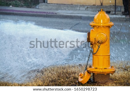 Yellow fire hydrant wide open gushing water onto the street with slightly grainy effect where water is falling back down over the pavement Royalty-Free Stock Photo #1650298723