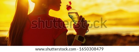Dehydrated runner fitness woman drinking water bottle sports drink on summer heat workout after run training jogging outdoors at sunset panoramic banner. Girl running silhouette against sun flare. Royalty-Free Stock Photo #1650308479