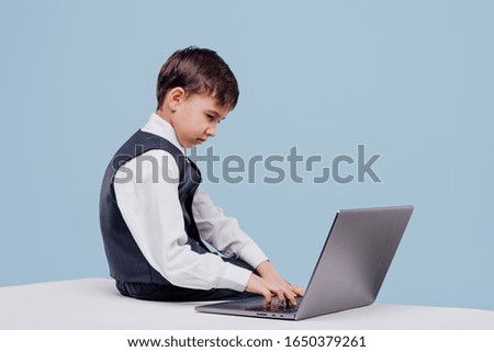 side view of curious little boy typing on laptop while sitting on table in studio on blue background