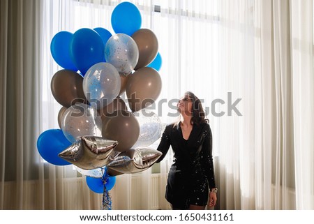 Young pretty woman with balloons indoors against the background of a large window with curtains. Balls are blue, white and golden. Festive surprise, birthday present.