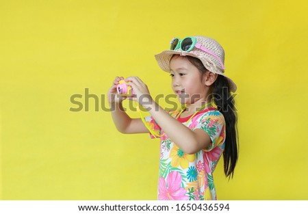 Attractive asian little girl in floral pattern summer dress and hat with sunglasses taking photo by toy camera isolated on yellow background. Fashion of Holiday and summer concept.