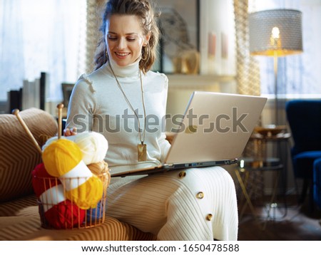 happy elegant middle age housewife in white sweater and skirt with basket with knitting yarn and needles learn how to knit using website on a laptop in the modern house in sunny winter day.