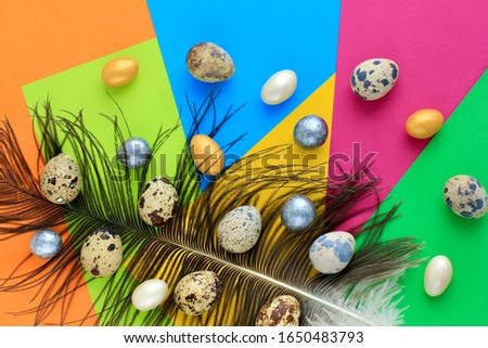 Quail eggs motley coloring, white, gold and silver candies lying on a large feather on bright colorful geometric background. Creative minimal trend concept of food, Easter. Copy space.