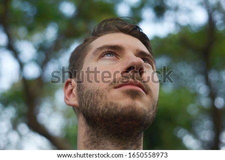 A caucasian bearded man face portrait. He gives 45 degree angle of face to the camera. His eyes look upward into the sky. There are trees in his background Royalty-Free Stock Photo #1650558973