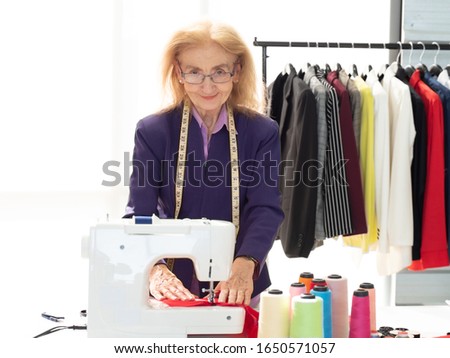 Senior woman standing at desk look at camera smiling while checking her work on sewing machine in the clothing factory. People, age, job and professional lifestyle. Copy space.