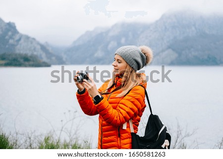 A beautiful blonde tourist woman, with a wool hat, backpack and orange coat, uses the camera in a landscape with a lake and mountains in the background