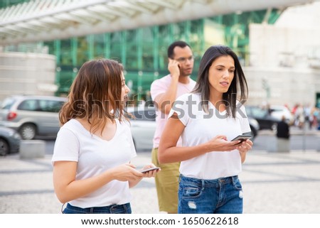 Two women walking on street with phones. Cheerful friends holding smartphones while strolling. Communication and technology concept