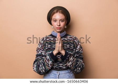 Photo of caucasian calm woman in earrings posing with holding palms together isolated over beige background