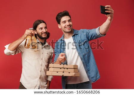 Two cheerful young men wearing casual clothes standing isolated over red background, showing pizza in boxes and bottles of beer, taking a selfie