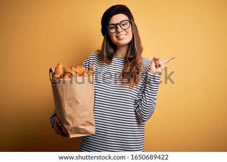 Young beautiful brunette french woman wearing beret holding bag with homemade croissants very happy pointing with hand and finger to the side