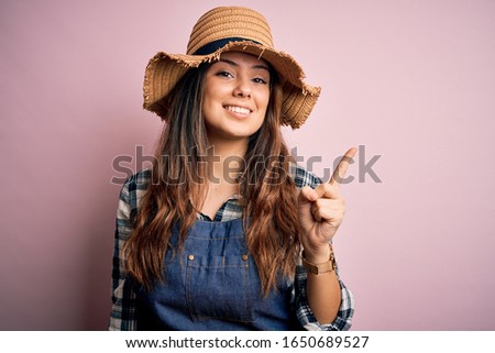 Young beautiful brunette farmer woman wearing apron and hat over pink background with a big smile on face, pointing with hand and finger to the side looking at the camera.