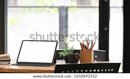 Photo of Computer tablet with white blank screen putting on wooden table together with Pencil holder, Coffee cup, Stack of books. Orderly and Comfortable workplace concept.