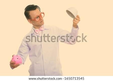 Studio shot of young crazy man doctor holding piggy bank and empty coffee cup upside down